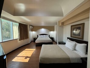 A hotel room with two beds, a large window with curtains, a framed artwork on the wall, and a ceiling light.