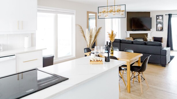 Een moderne keuken met een inbouwfornuis, een eethoek met een houten tafel en stoelen, een grijze bank en een televisie boven een open haard.
