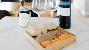 Een eettafel met een fles wijn, twee glaasjes, een potje room, en een houten dienblad met kaas en brood.