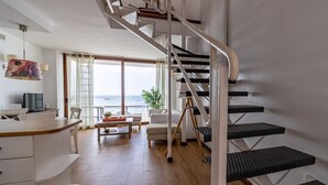 Un soggiorno moderno con pavimento in legno, arredi bianchi, una scala con gradini neri e vista sul mare attraverso ampie finestre.