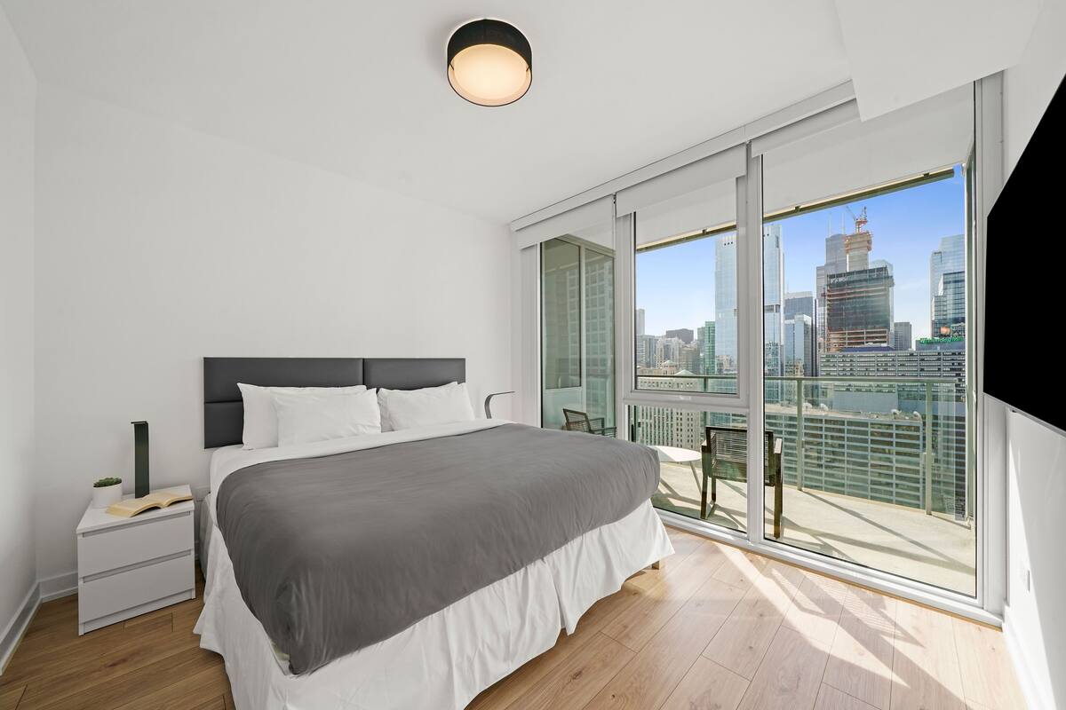 Une chambre à coucher moderne avec un grand lit, une table de chevet, une commode et une vue sur le paysage urbain grâce à de grandes fenêtres du sol au plafond.