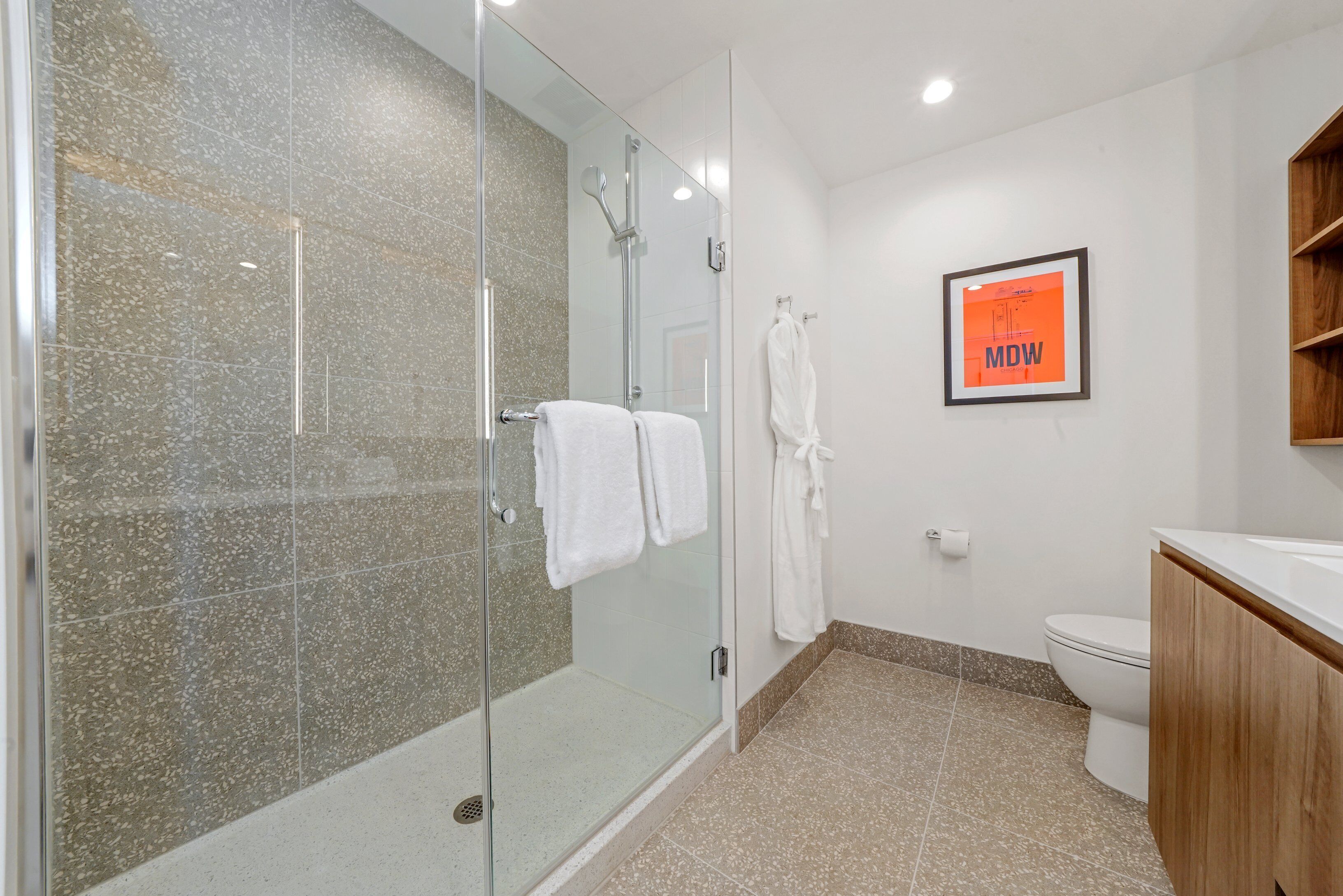 A modern bathroom with a glass shower enclosure, white tiled walls, a wooden vanity, and a framed picture on the wall.
