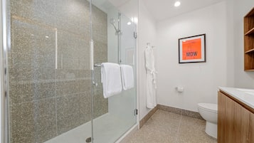 A modern bathroom with a glass shower enclosure, white tiled walls, a wooden vanity, and a framed picture on the wall.
