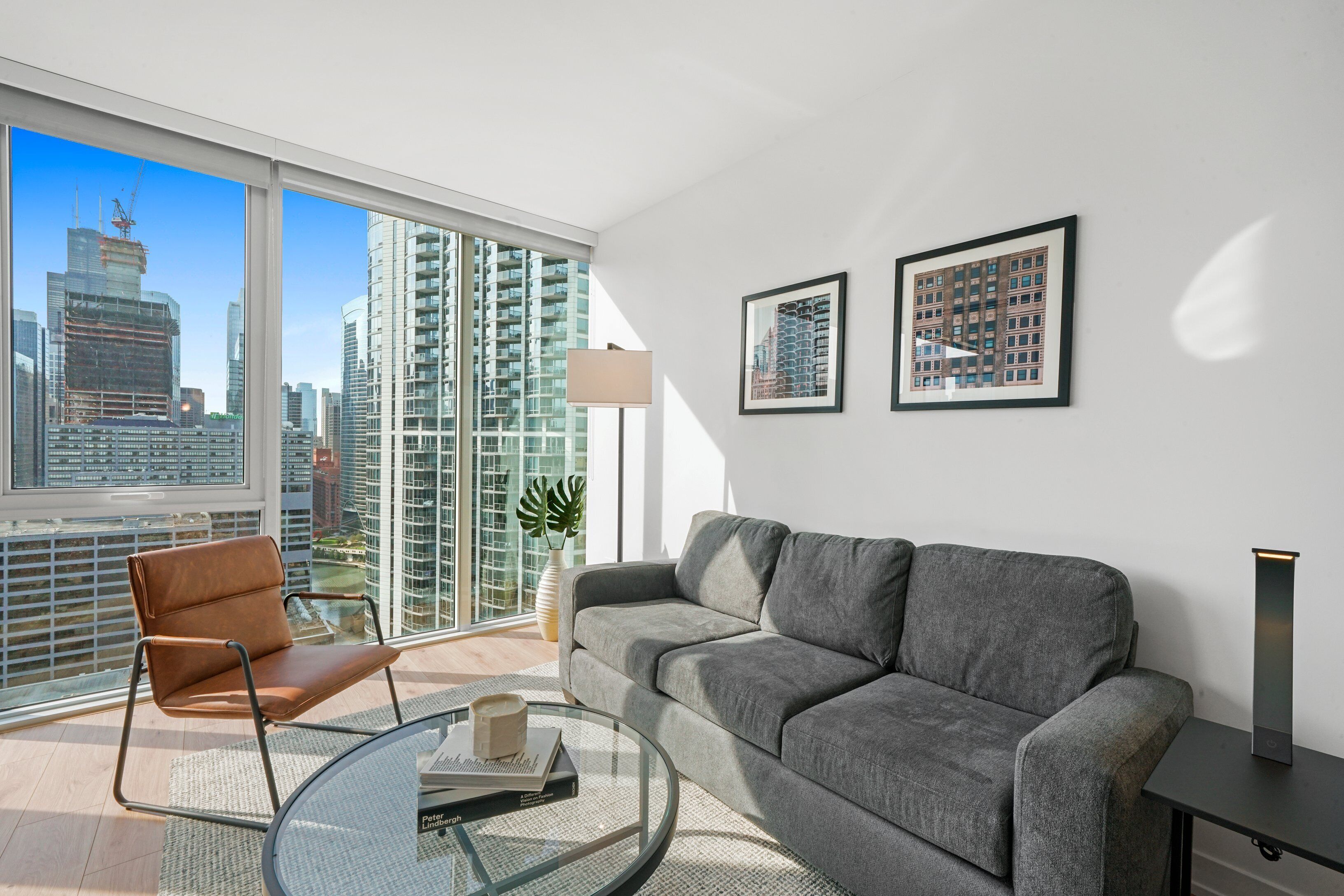 A modern living room with a grey sofa, a brown leather chair, a glass coffee table, and a city view through large windows.