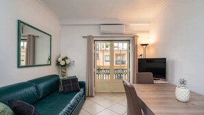 A living room with a green sofa, a wooden dining table, a TV, and a balcony with a view of buildings. - Apartment for 4 with privileged access to the beach (Albufeira)