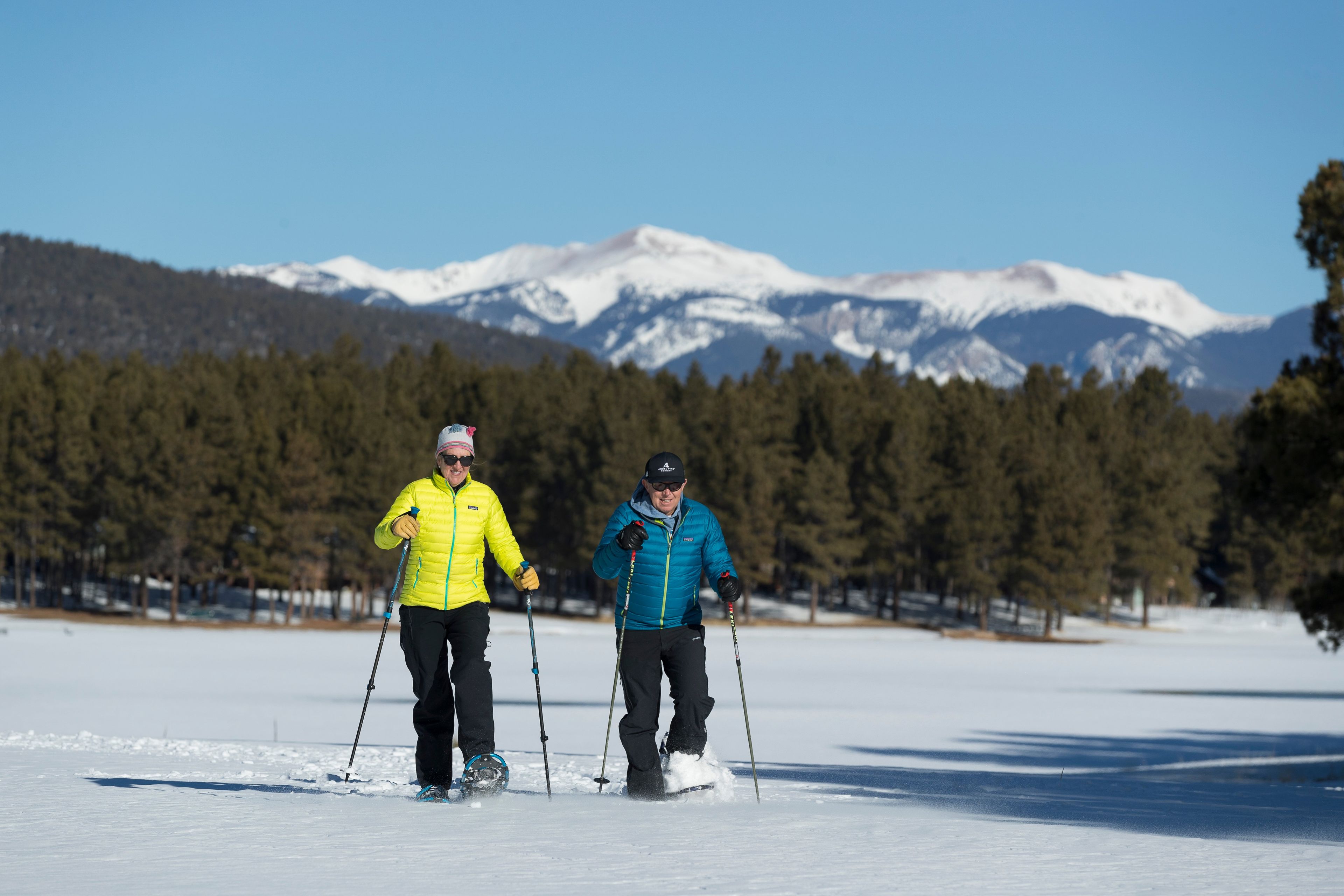 Dues persones practiquen esquí de fons sobre un camp nevat amb una serralada de muntanyes al fons.