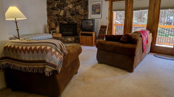 A cozy living room with a stone fireplace, a television, and a large window with a view of the outdoors. - Cozy Condo close to the Chair Lift (Angel Fire)