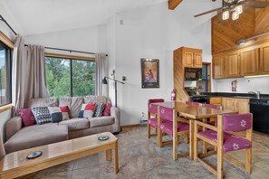 A modern living room with a sofa, a coffee table, and a dining table with chairs. There is a kitchen area with wooden cabinets and a window.