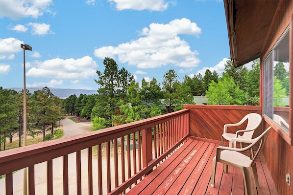 A wooden deck with a red railing, a white plastic chair, and a view of a green landscape with trees and a mountain in the distance.
