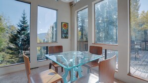 Un comedor con mesa de vidrio y sillas de madera, ventanas amplias con vistas a árboles y montañas, y una lámpara de techo.