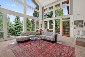 A spacious living room with a large window, a sectional sofa, a glass coffee table, and a red patterned rug.