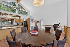 A spacious dining area with a round wooden table surrounded by chairs, a kitchen with wooden cabinets, and a view of trees through large windows.