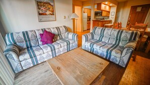 Una sala de estar con dos sofás estampados, una mesa de centro de madera y una cocina al fondo.