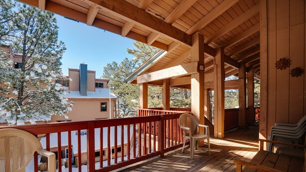 A wooden deck with a red railing, chairs, and a view of a building with snow-covered trees.