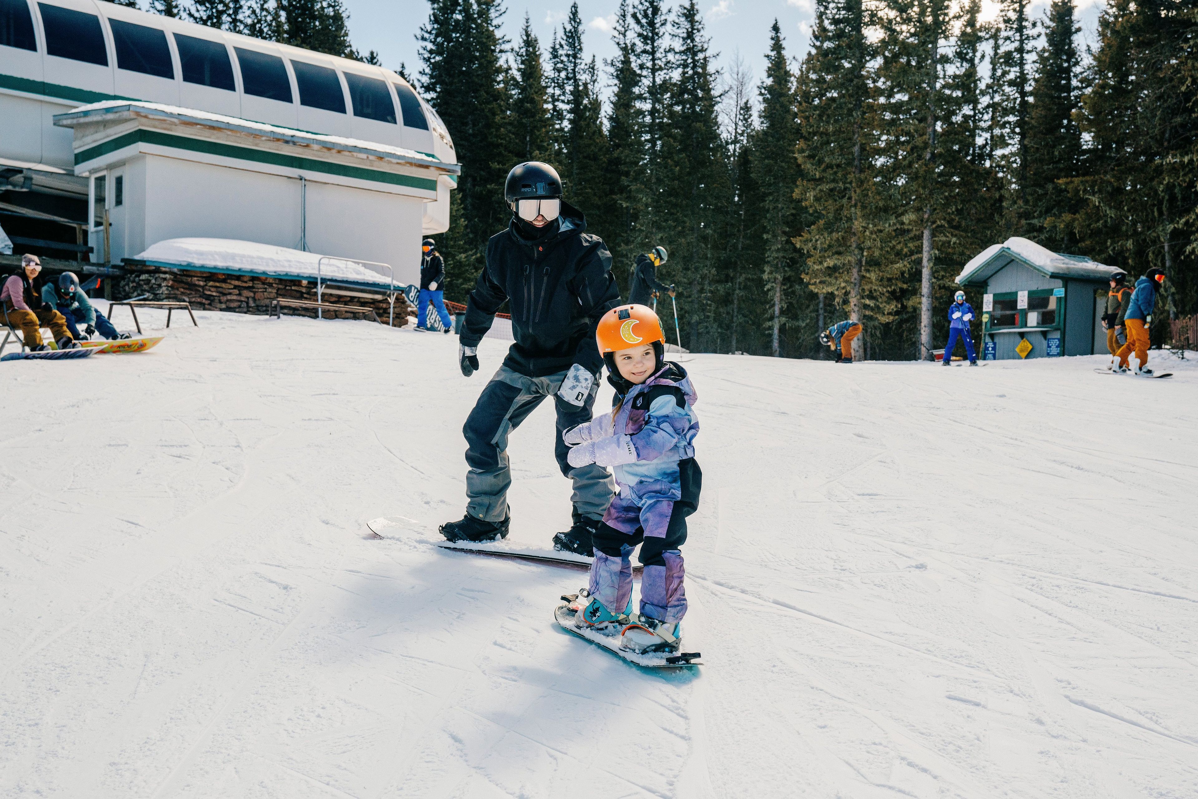 A snowy ski slope with a person and a child snowboarding.
