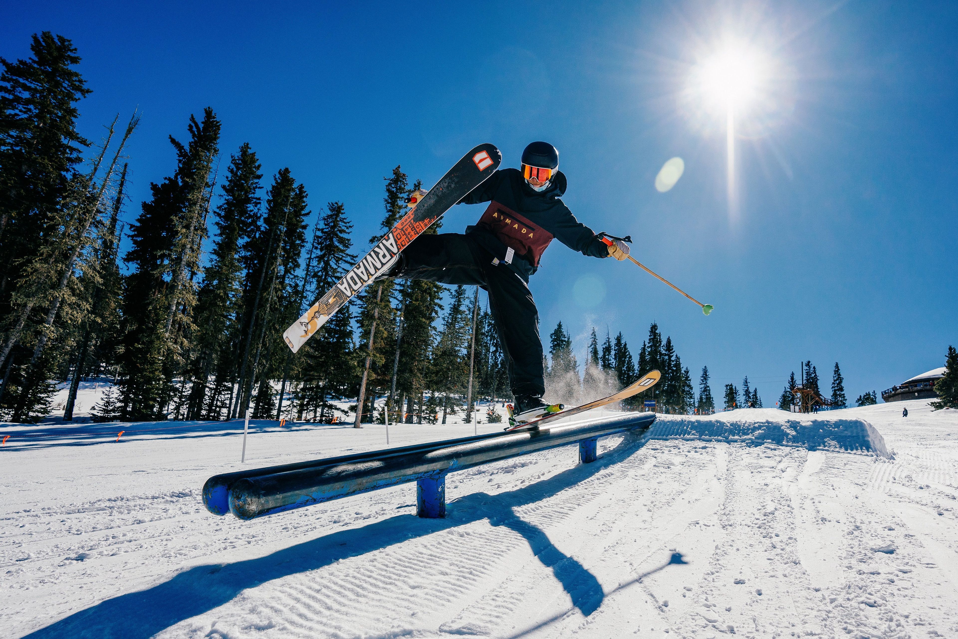 Ein Skifahrer führt einen Rail-Grind-Trick auf einem verschneiten Hang aus, im Hintergrund sind Bäume zu sehen.