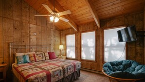 Un dormitorio de cabaña de madera con una cama, un ventilador de techo, un televisor y una ventana con persianas.