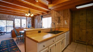 Interior de madera con una zona de cocina que incluye encimera de madera, lavamanos y armarios. Hay una zona de comedor con una mesa y sillas, y un ventanal que proporciona luz natural.