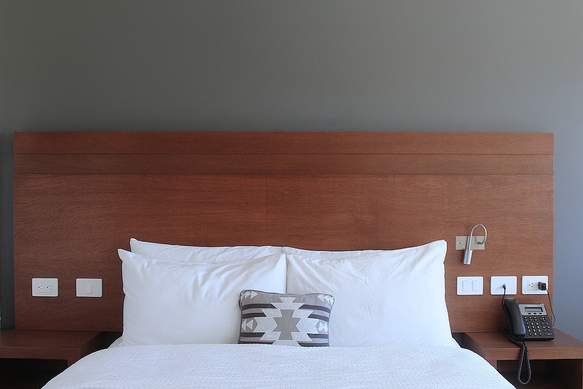 Une chambre d’hôtel avec une tête de lit en bois, du linge de lit blanc, un coussin à motifs et un téléphone sur la table de chevet.
