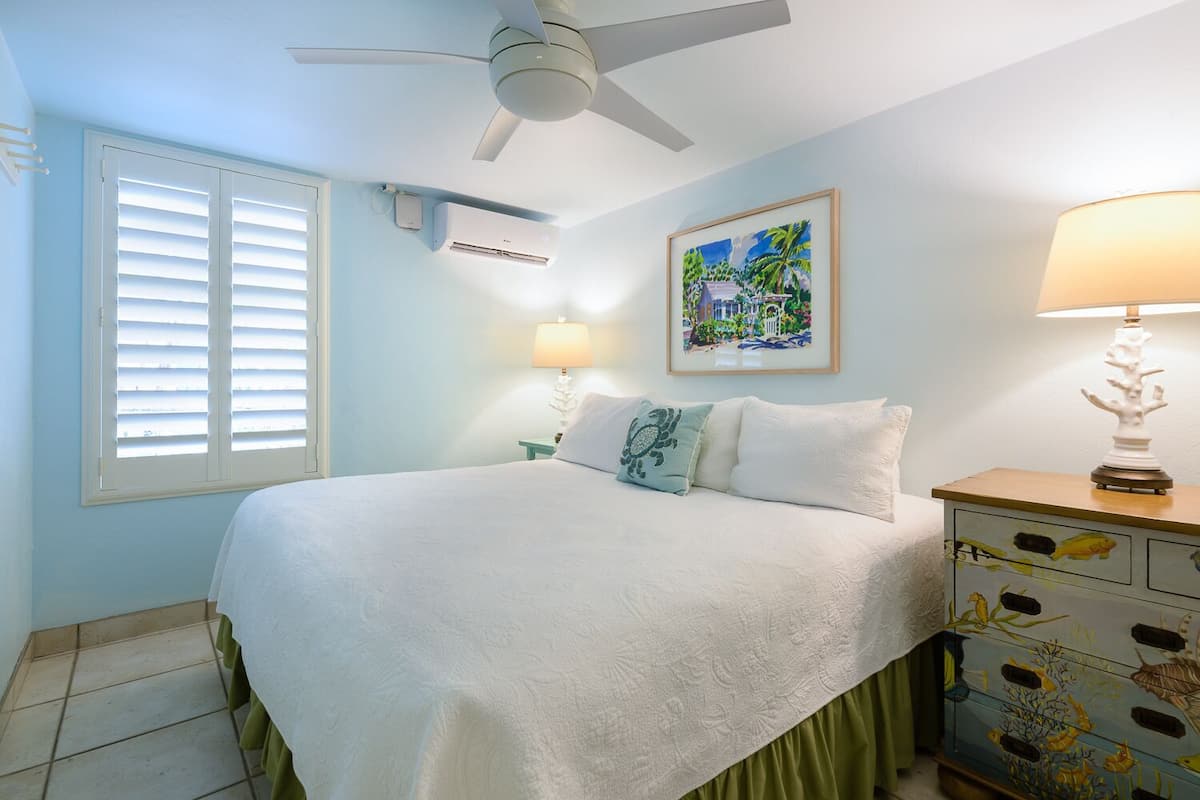 Une chambre à coucher comprenant un lit, un ventilateur de plafond, un appareil de climatisation, une table de chevet avec une lampe, une commode avec des tiroirs et une fenêtre avec des stores.