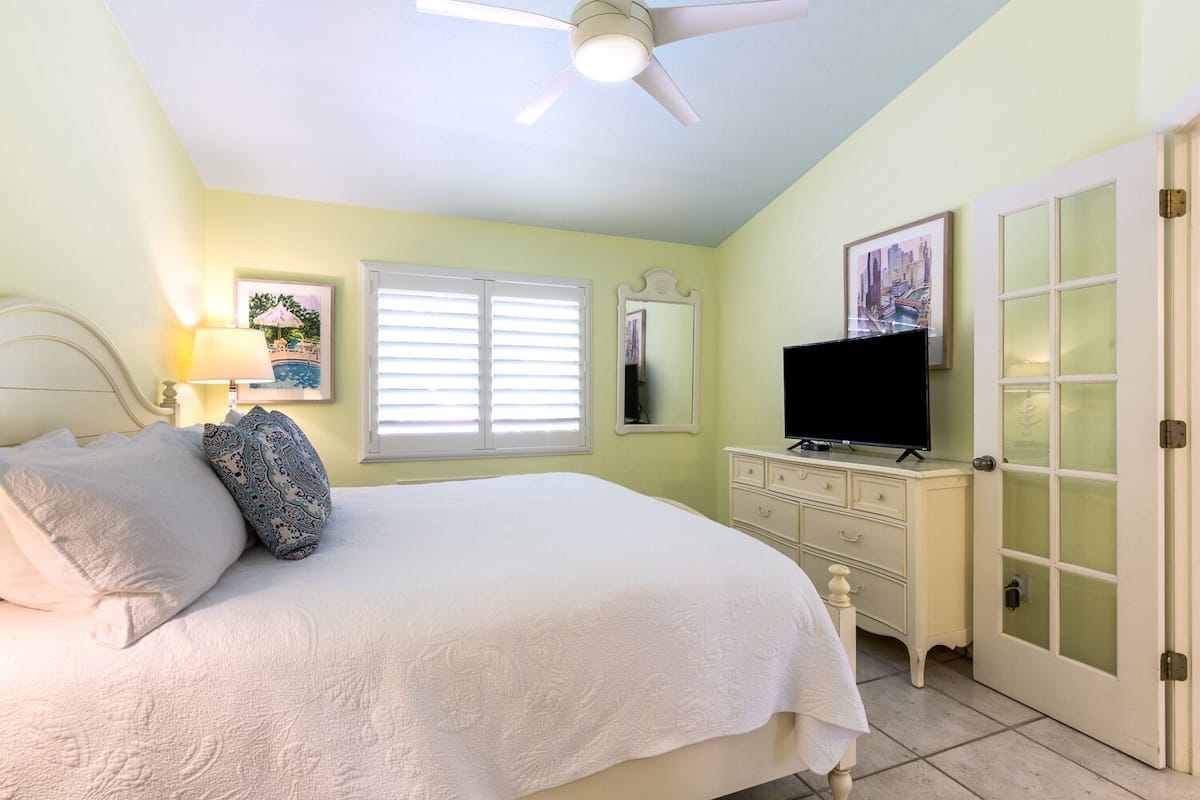 Une chambre à coucher comprenant un lit, une commode, un téléviseur, une fenêtre avec des stores, un ventilateur de plafond et une porte avec des panneaux en verre dépoli.