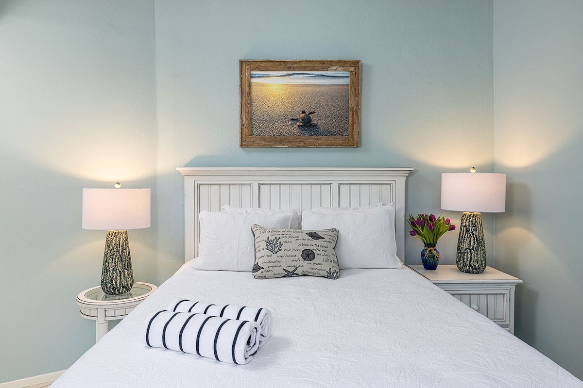 Un lit bien fait, recouvert d’une couette blanche et agrémenté d’oreillers à rayures, est encadré par deux tables de chevet sur lesquelles reposent des lampes et des vases décoratifs. Un tableau représentant une scène de plage est accroché au-dessus du lit.