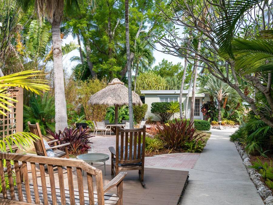 A tropical garden with wooden furniture, a thatched umbrella, and a pathway leading to a building.