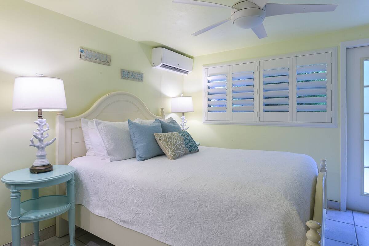 Une chambre à coucher comprenant un lit, deux tables de chevet, un ventilateur de plafond et une fenêtre avec des volets.