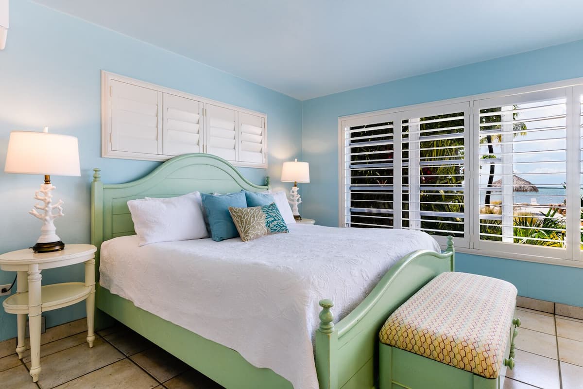 Une chambre à coucher comprenant un lit, deux tables de chevet, une fenêtre avec des stores et une vue sur l’extérieur.