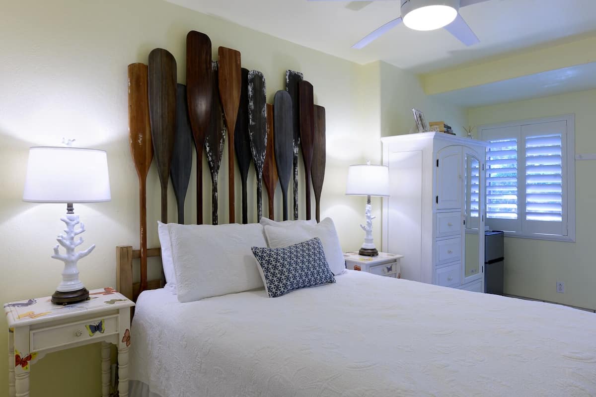 Une chambre à coucher comprenant un lit, deux tables de chevet, un ventilateur de plafond, une armoire et une fenêtre avec des stores.