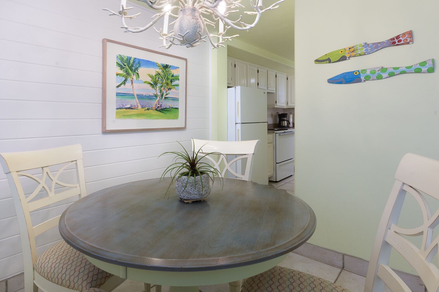Un coin repas avec une table ronde, deux chaises, une plante en pot, un tableau représentant une scène de plage et, au fond, une cuisine.