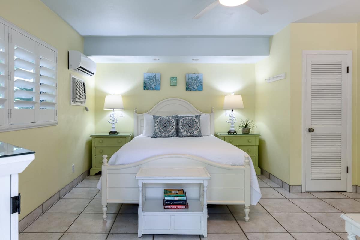 Une chambre à coucher comprenant un lit, deux tables de chevet, une commode, une petite table avec des livres et une porte.