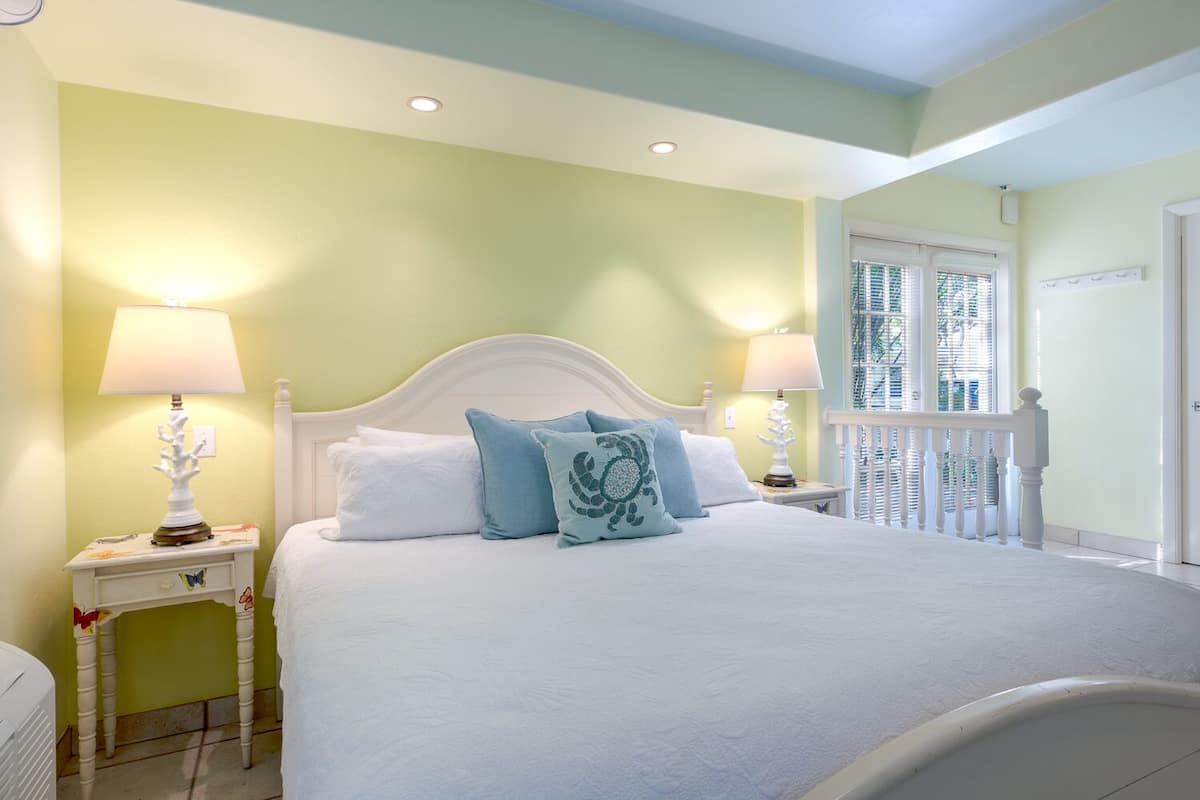 Une chambre avec un lit blanc, deux tables de chevet avec des lampes, une fenêtre avec des stores et une rambarde de balcon.