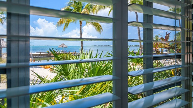 A view through a window with metal bars, featuring a beach with palm trees, a thatched umbrella, and a wooden dock. - Kona Kai Resort and Gallery - Adults Only (Key Largo)