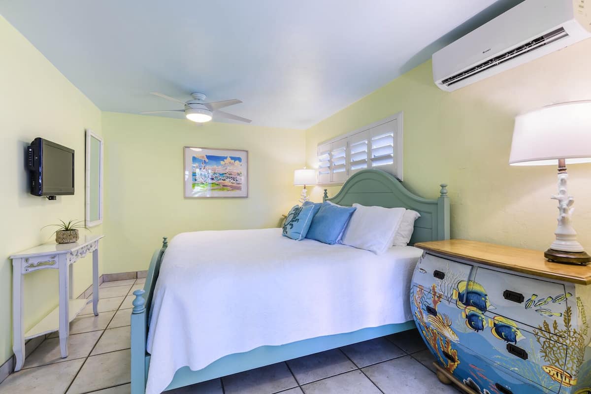 Une chambre à coucher comprenant un lit, un ventilateur de plafond, un climatiseur, une télévision, un tableau encadré, une table de chevet et une commode décorée de motifs de poissons.