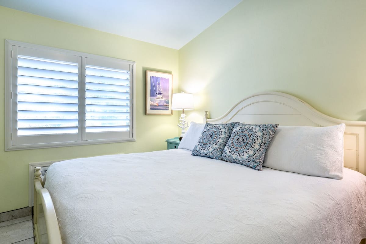 Une chambre à coucher avec un lit, deux oreillers, une table de chevet, une lampe et une fenêtre avec des stores.