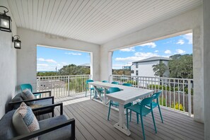 A covered outdoor deck with a white table and chairs, a sofa with patterned cushions, and a view of neighboring houses and trees.