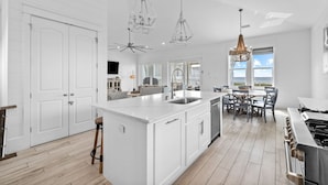 Une cuisine spacieuse dotée d’un grand îlot central, de meubles blancs et d’une cuisinière à gaz. On y trouve des luminaires suspendus et un lustre.