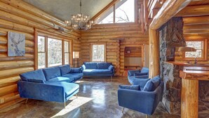 A log cabin interior with a sectional sofa, a stone fireplace, and large windows. - Custom Black Hills Log Home w/views, Secluded, Only 12 miles to Mt Rushmore! (Hermosa)