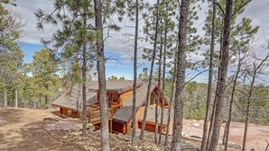 A log cabin with a deck and large windows, surrounded by tall pine trees. - Custom Black Hills Log Home w/views, Secluded, Only 12 miles to Mt Rushmore! (Hermosa)