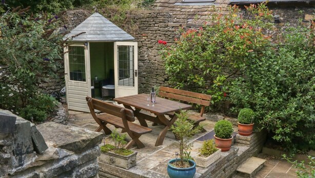 Une terrasse en pierre avec une table et des bancs en bois, entourée de plantes en pot et d’un petit abri de jardin.