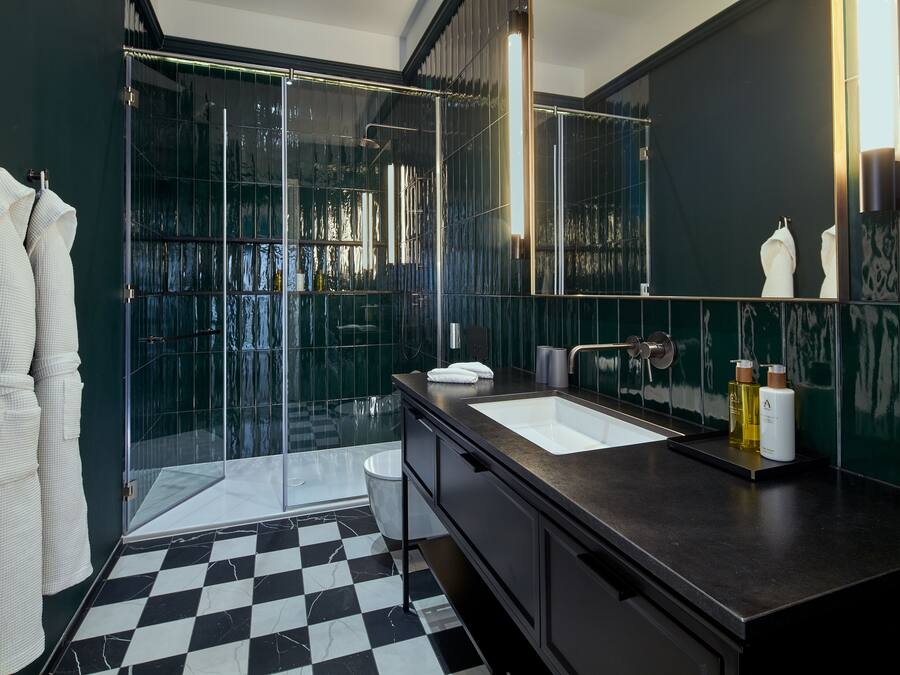 Un baño moderno con piso a cuadros blanco y negro, cabina de ducha de vidrio, espejo grande y encimera oscura con lavamanos y artículos de aseo.