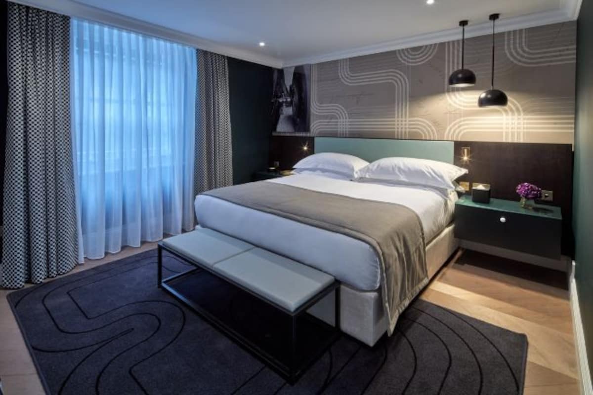 Habitación de hotel moderna con una cama grande, mesitas de noche, un banco y vistas al exterior.