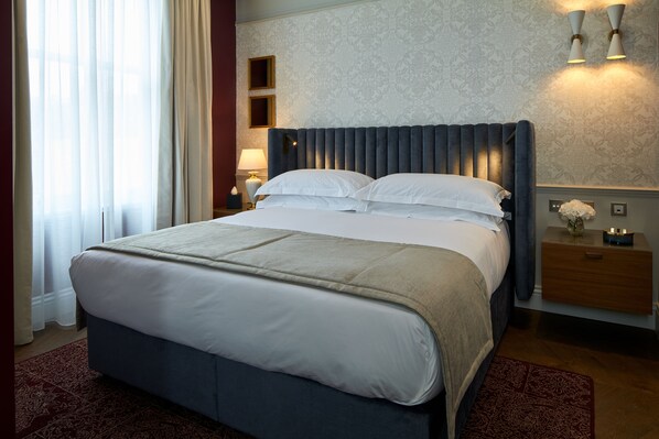 A neatly made bed with a grey headboard, a bedside table with a lamp, and a floral-patterned wall. - Lexham Gardens by Cheval Maison (London)