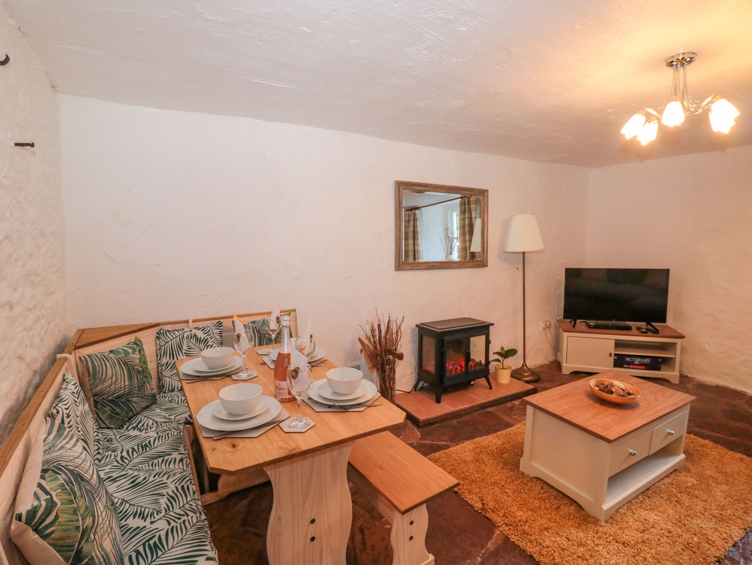 A cozy living room with a wooden dining table set for four, a sofa with patterned cushions, a television on a wooden stand, and a fireplace.