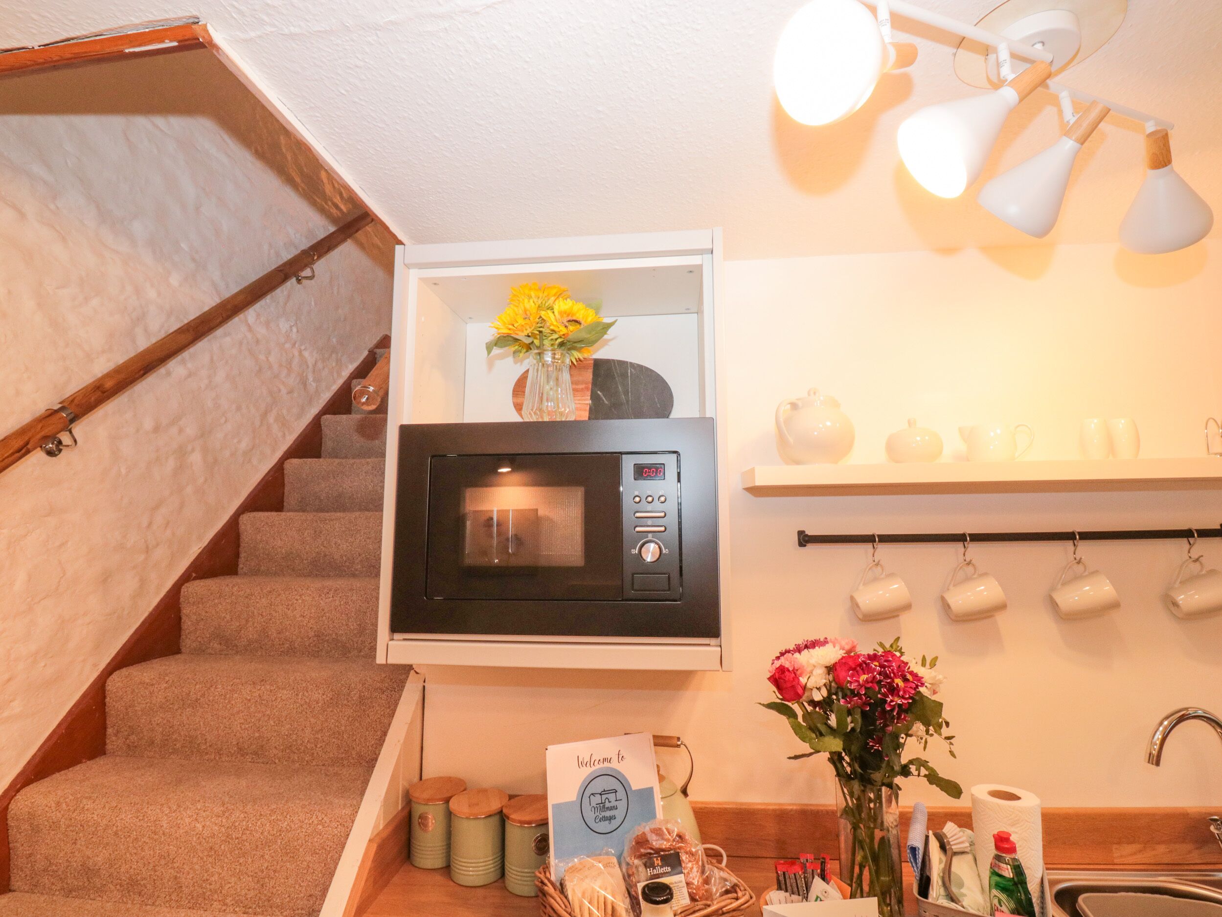 A kitchen area with a microwave, a shelf with white mugs, a vase with flowers, and a welcome sign.