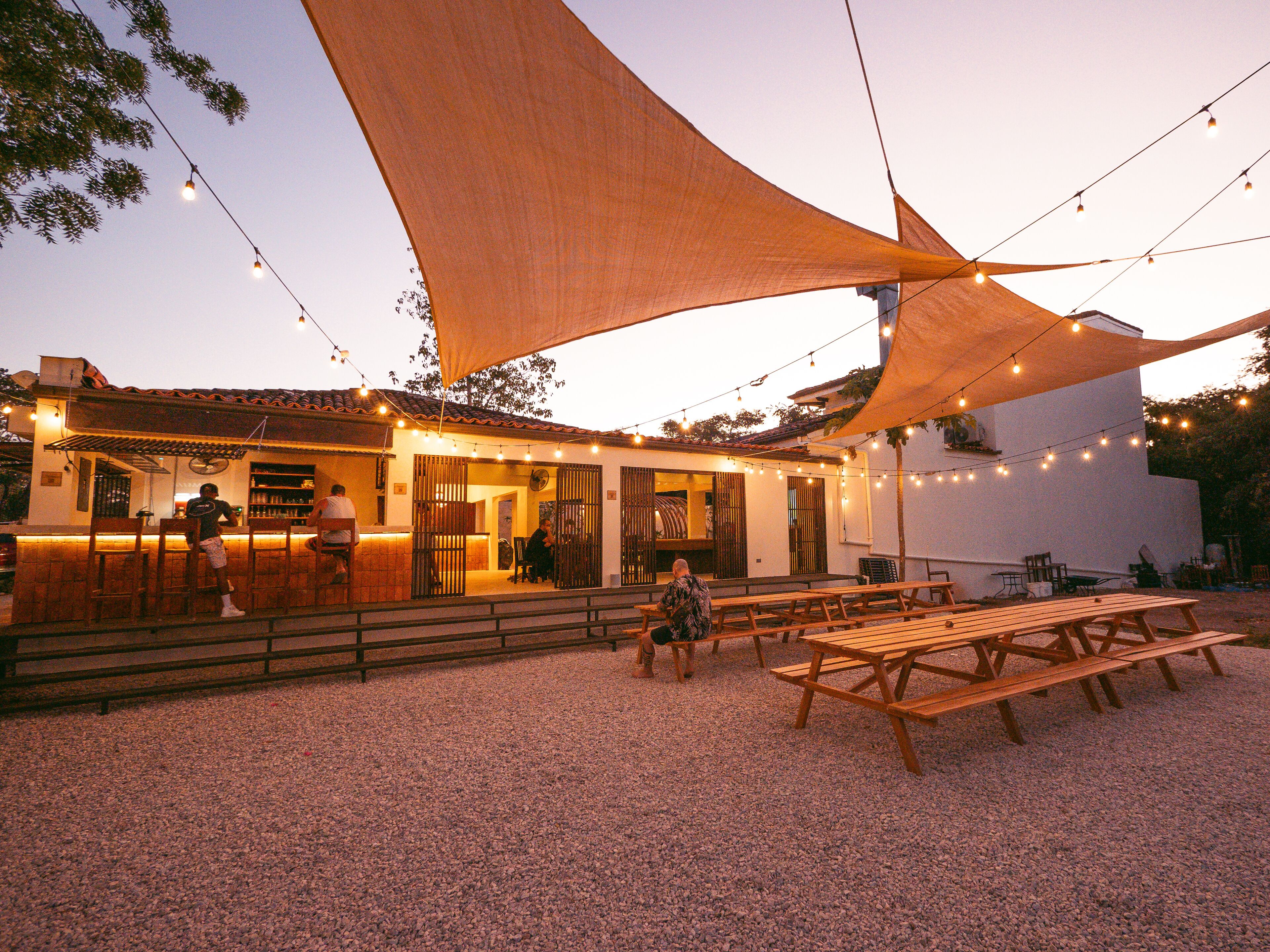 An outdoor seating area with wooden tables and benches, string lights, and a large shade sail.