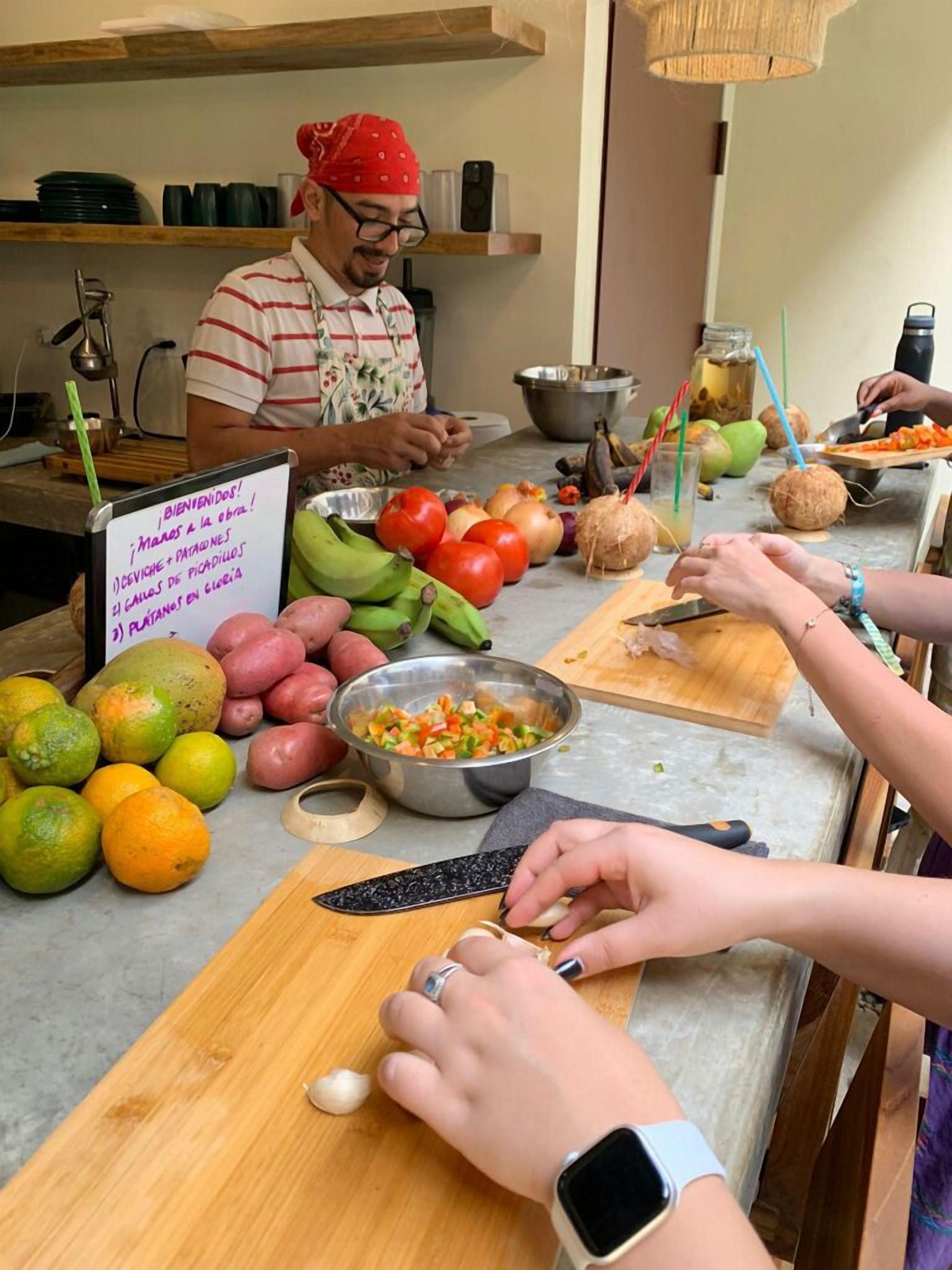 Una cocina donde una persona prepara alimentos, con diversas frutas y verduras sobre la encimera, y un cartel con texto en español.