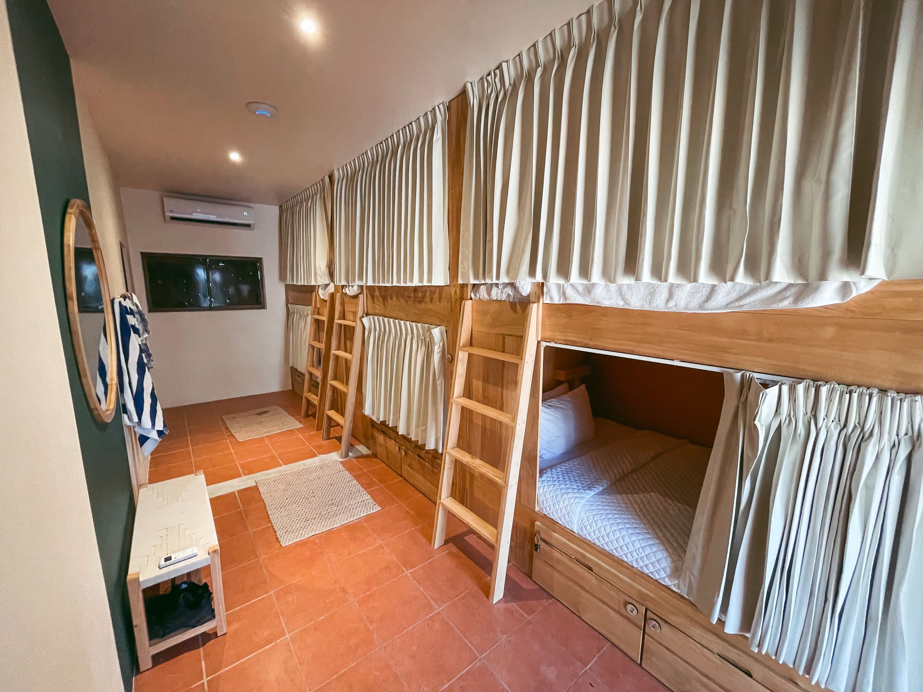 Quarto tipo dormitório com beliches, uma escrivaninha pequena, um espelho e uma janela.
