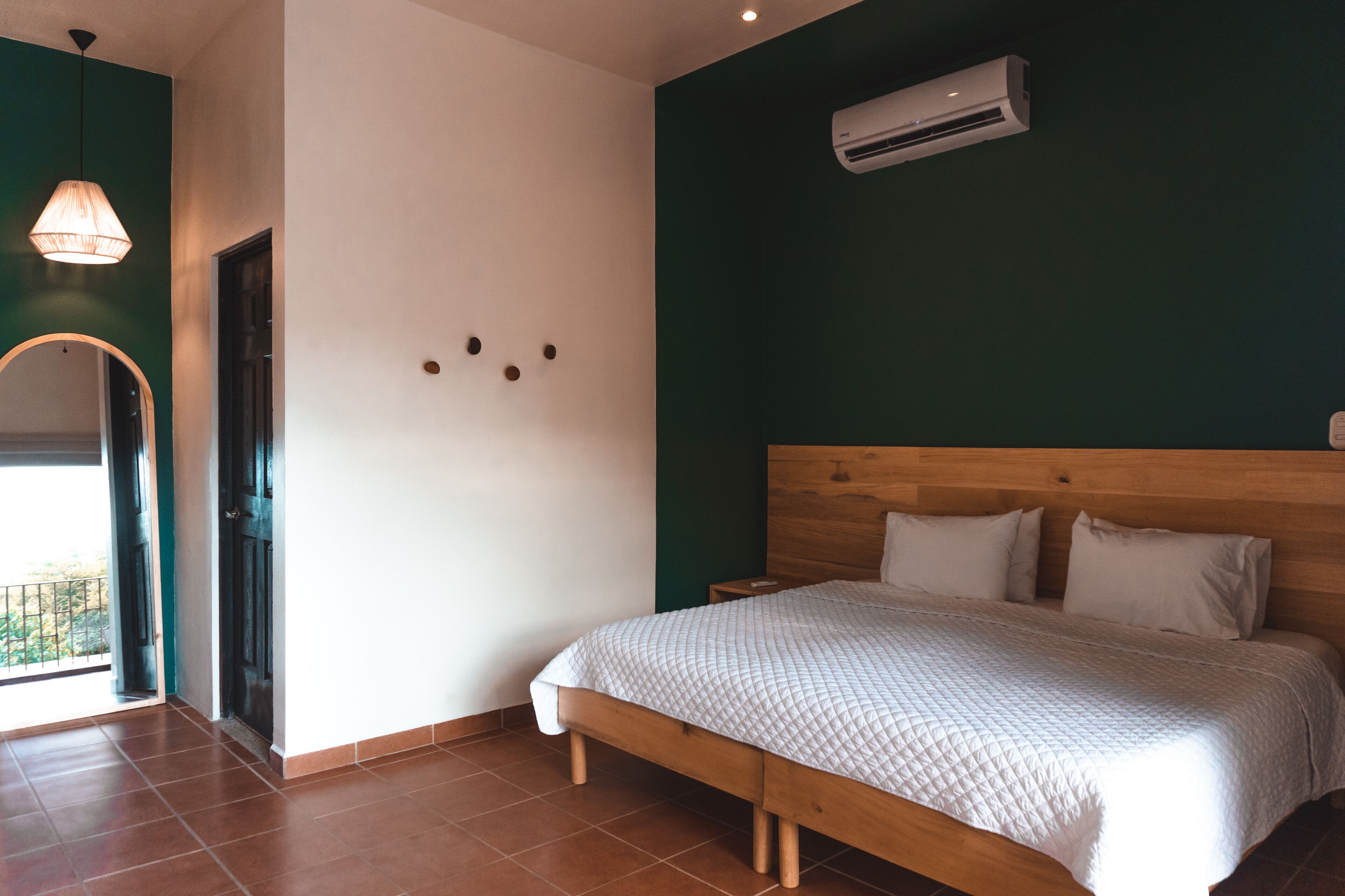A bedroom with a wooden bed, a white quilted bedspread, a wooden headboard, a wall-mounted air conditioner, and a balcony door.